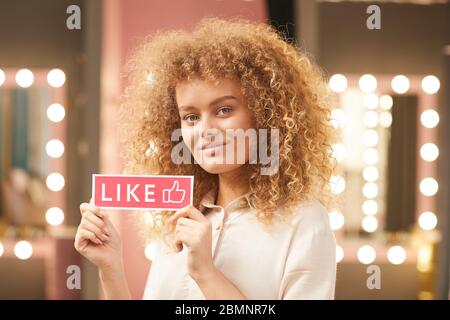 Portrait of modern curly-haired woman holding LIKE sign while posing in glamorous dressing room interior, copy space Stock Photo