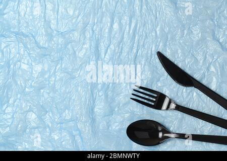 Disposable black appliances fork, spoon, knife on blue crumpled polyethylene. Eco, zero waste concept. Flat lay, top view. Horizontal. Stock Photo