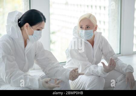 Two female medical workers in protective clothing and medical mask sitting next to the window, talking Stock Photo