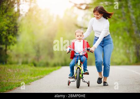 Mom teaches little son to ride bike in park, keep balance, have fun family. Mother day concept. Stock Photo