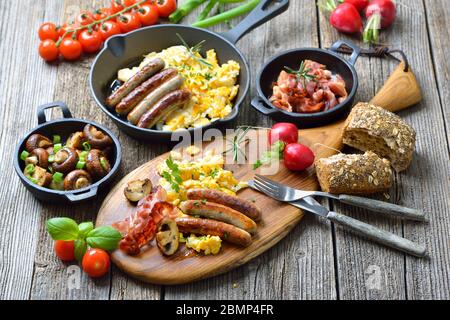 A hearty breakfast with scrambled eggs with bacon, Nuremberg sausages and fried mushrooms on a rustic wooden table Stock Photo