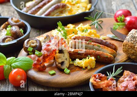 A hearty breakfast with scrambled eggs with bacon, Nuremberg sausages and fried mushrooms on a rustic wooden table Stock Photo