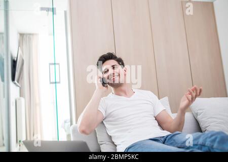 Image of a cheery pleased young man indoors at home on sofa using laptop computer talking by mobile phone. Stock Photo