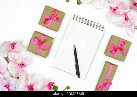 flatlay of Gift present boxes with pink ribbon and orchid flowers on white background top view. Note pad writing. Stock Photo