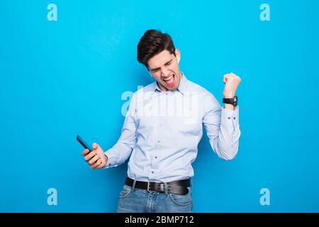 Image of a handsome excited young man using mobile phone isolated over blue wall background make winner gesture. Stock Photo