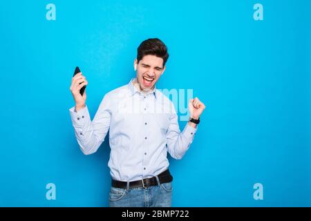 Image of a handsome excited young man using mobile phone isolated over blue wall background make winner gesture. Stock Photo