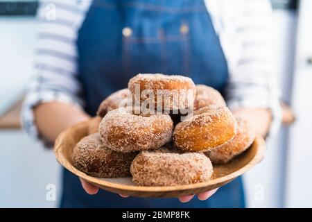 Close up of a young woman in denim apron holding a plate of freshly baked homemade donuts with sugar ready for sale Stock Photo