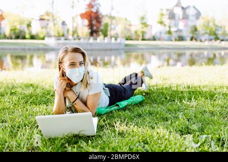 Girl working at the computer lying on the grass in a protective medical mask on her face. Freelance, quarantine concept. Stock Photo