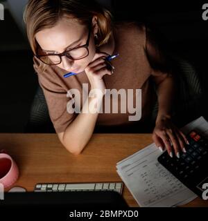 Woman in glasses using calculator to calculate the invoice, planning expenses, holding a pen near face, working on desktop pc late at night at home of Stock Photo