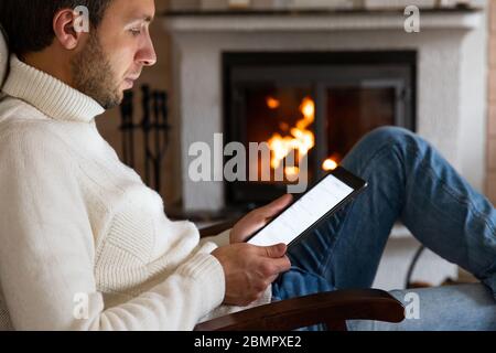 Close up of caucasian man wear high neck white sweater using a digital tablet, reading news, scrolling social networks, rested in a living room by the Stock Photo