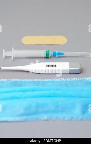 Vertical image of surgical mask, syringe with needle and digital thermometer with adhesive plaster on gray background. Protection against coronavirus. Stock Photo