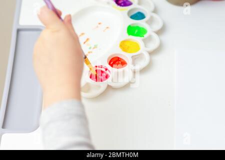 The child is ready to paint with watercolors. Child's hand holding brush over palette on blank sheet of white paper with watercolors. Stock Photo