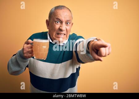 Senior handsome man drinking jar of beer standing over isolated yellow background pointing displeased and frustrated to the camera, angry and furious Stock Photo