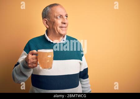 Senior handsome man drinking jar of beer standing over isolated yellow background smiling looking to the side and staring away thinking. Stock Photo