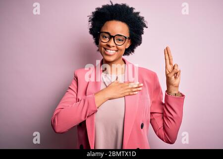 Young beautiful African American afro businesswoman with curly hair wearing pink jacket smiling swearing with hand on chest and fingers up, making a l Stock Photo
