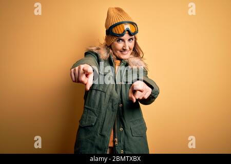 Middle age beautiful blonde skier woman wearing snow sportwear and ski goggles pointing to you and the camera with fingers, smiling positive and cheer Stock Photo