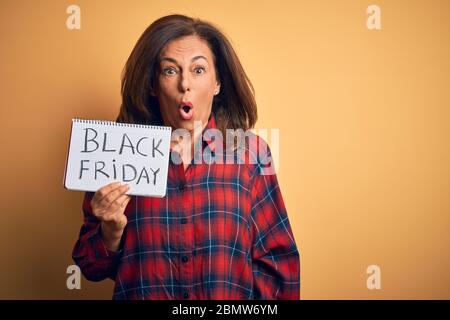 Middle age brunette woman holding black friday paper ad over isolated background scared in shock with a surprise face, afraid and excited with fear ex Stock Photo