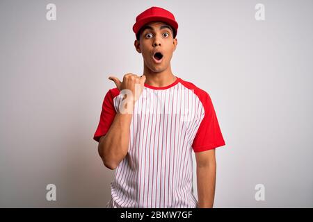 Young handsome african american sportsman wearing striped baseball t-shirt and cap Surprised pointing with hand finger to the side, open mouth amazed Stock Photo