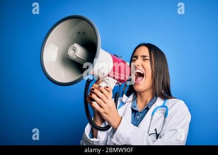 Hispanic doctor woman wearing medical white coat shouting angry on protest through megaphone. Yelling excited on ludspeaker talking and screaming news Stock Photo