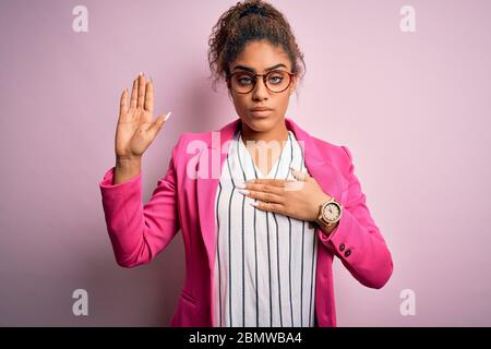 Beautiful african american businesswoman wearing jacket and glasses over pink background Swearing with hand on chest and open palm, making a loyalty p Stock Photo