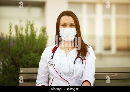 Young female doctor in surgical mask and earbuds during online webinar or conference outdoors via wireless internet Stock Photo