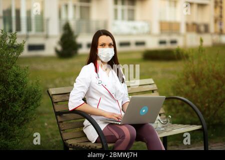 Female medical doctor in mask and stethoscope uses laptop and looks at you Stock Photo