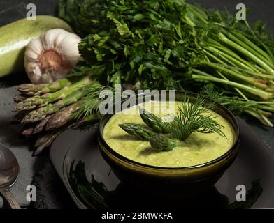 Purred creamy asparagus soup in glass bowl on black plate against raw fresh asparagus and greenery. Stock Photo