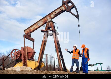 Two oil engineers wearing overalls, orange vests, and helmets, standing on an oilfield next to oil pump jack discussing something on a sunny day. Concept of petroleum industry and oil extraction. Stock Photo