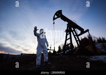 Silhouette of male scientist showing stop sign while standing on territory of oil field with pump jack and beautiful sky on background. Environmentalist wearing white radiation suit, gas mask, gloves Stock Photo