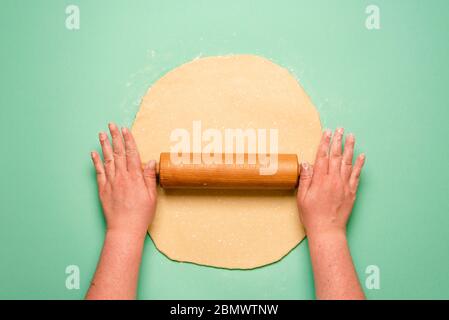 Woman's hands stretching pie dough with a wooden rolling pin. Top view with dough on a green kitchen counter. Stock Photo