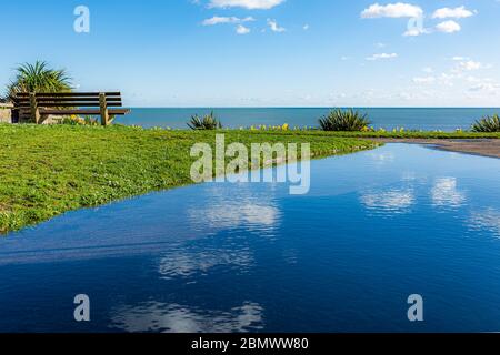 Landscape photograph of blue sky reflected in large puddle on public viewpoint overlooking sea Stock Photo