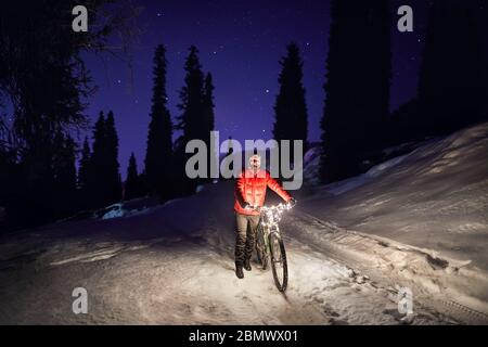 Man in red jacket with bicycle at winter snowy forest in the mountains under night sky with stars