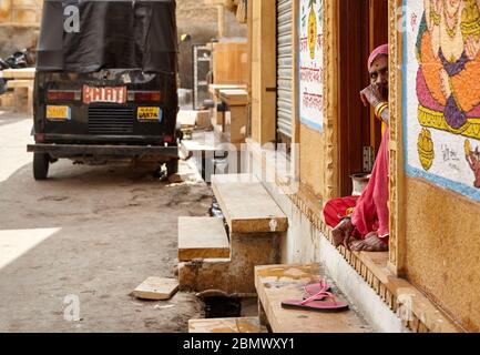 JAISALMER, INDIA – MARCH 14, 2015: Old Indian woman in pink clothes is sitting at the stairs at her house with Hindu paintings in Jaisalmer city, Indi Stock Photo