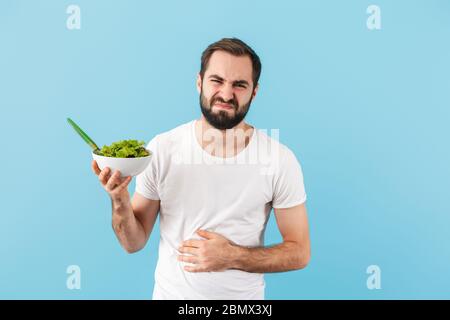 Handsome disgusted young bearded man wearing t-shirt standing isolated over blue background, suffering from a stomach cramp Stock Photo