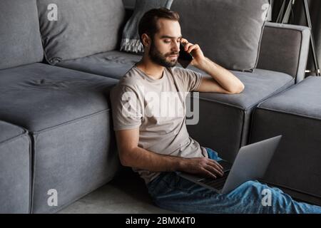 Image of a concentrated young man indoors at home on sofa using laptop computer talking by mobile phone. Stock Photo