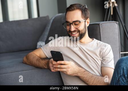 Attractive smart young man sitting on a floor in the living room, using mobile phone Stock Photo