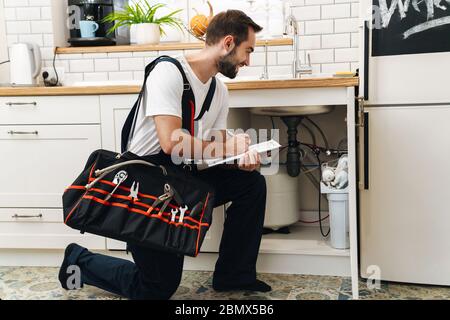 Image of young caucasian plumber man in uniform holding bag with equipment and clipboard while working in apartment Stock Photo