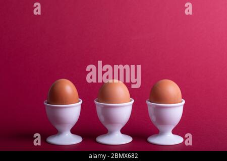 three brown eggs in white eggs cups before red background, close-up Stock Photo