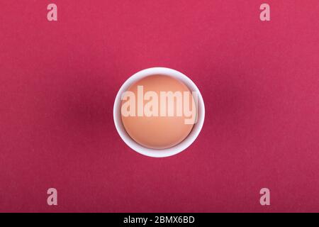 one brown egg in white egg cup before red background, close-up Stock Photo