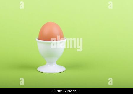 one brown egg in white egg cup before green background, close-up Stock Photo