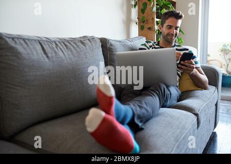 Smiling man using a cellphone and laptop on his sofa Stock Photo