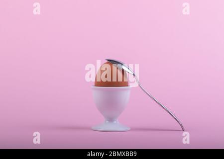 one brown egg in white egg cup before pink background, a teaspoon is laying on it, close-up Stock Photo