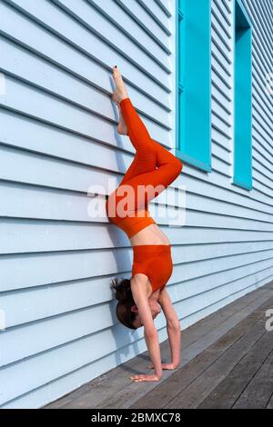 woman wearing orange sportswear doing a handstand against a blue wall. Stock Photo