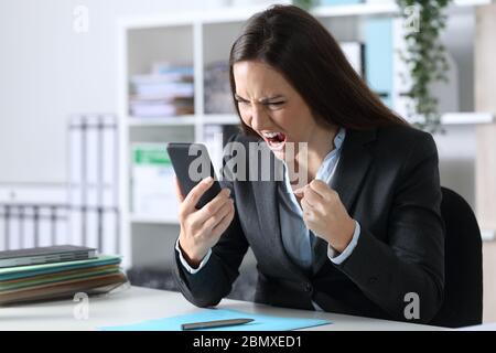 Angry executive woman reading bad news on smart phone sitting on a desk at office Stock Photo