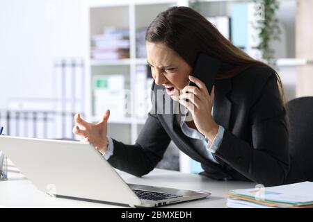 Angry executive woman calling on smart phone looking at laptop sitting on her desk at office Stock Photo