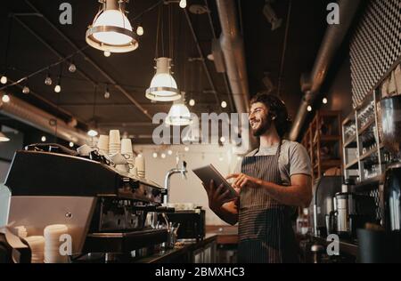 Smiling caucasian man wearing apron standing behind cafe counter using digital tablet looking away Stock Photo