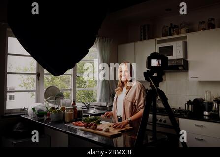 Cheerful female food blogger recording video for vlog at camera while standing in modern kitchen and chopping vegetables - vlogger vlogging from home Stock Photo
