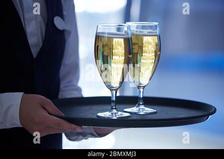 The hands of a waiter dressed in a black vest and a white shirt holding two glasses of champagne on a tray Stock Photo