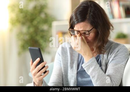 Adult woman suffers eyestrain with eyeglasses and holding phone sitting on a couch at home Stock Photo