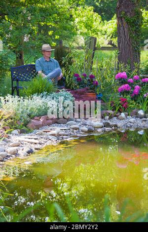 Mature man sitting on a bench by a pond in a garden with colorful flowers - focus on the man Stock Photo